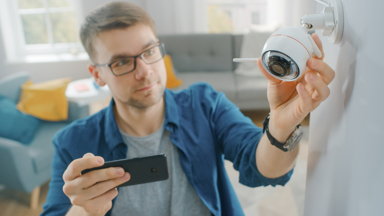 Young Man Adjusting a Modern Wi-Fi Surveillance Camera with Two Antennas on a White Wall at Home. He's Checking the Video Feed on his Smartphone.