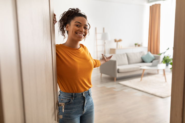 Cheerful Woman Opening Door And Gesturing Welcoming You To Come In