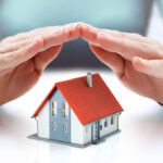 a shot that is used as the featured image of an article about home warranties. A person holding his hands protecting a small model of a home on a table